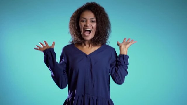 Surprised excited happy african woman jumping with ecstatic face expressions. Winning young female model on blue background.