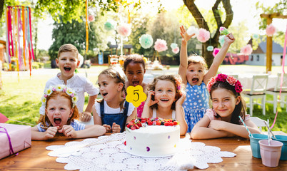 Children with cake standing around table on birthday party in garden in summer. - obrazy, fototapety, plakaty