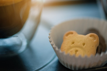 Hot espresso coffee in a clear glass, placed on a metal tray and wooden layer beside the coffee cup with a bear shaped cookie . All laid on dark brown wooden tables in retro coffee shop.
