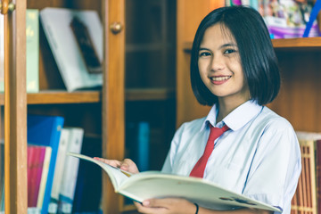 Asian high school female students with short hair in a white school uniform, red necktie, studying in the school library that made of wall-mounted bookcase with wood patterns.