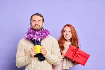 Playful joyful young lady with red curly hair, standing behind wide back of handsome male, preparing Christmas surprise, expressing cheerfulness, expecting positive emotions