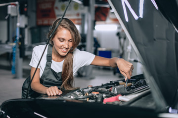 A brunette in a black jumpsuit and a white t-shirt near the open hood of black car. Young female in the garage is smiling at the camera and lowered gaze. car repair concept