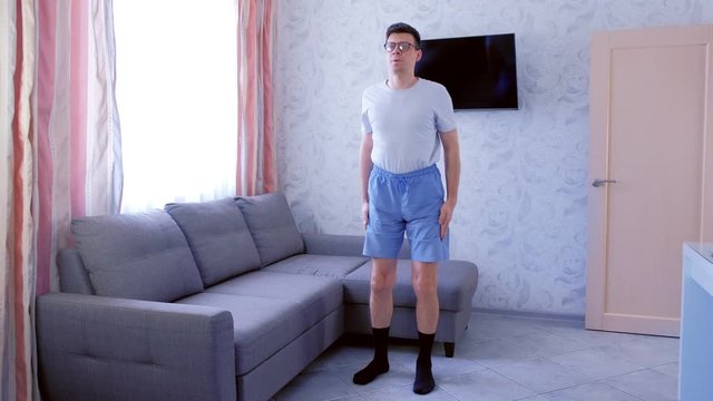 Funny nerd man in glasses and shorts without sportswear is making squats exercise at home. Funny pulls up shorts before exercise. Front view.
