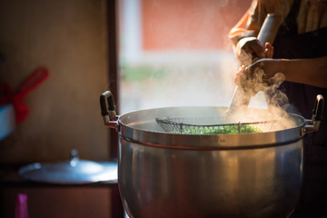 Cooking person in the restaurant is cooking while using a sieve for scalding green noodles in a large pot. And the water is boiling,