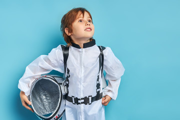 Caucasian child in white protective suit hold helmet in hands, surprisingly looking side at stars. Isolated blue background