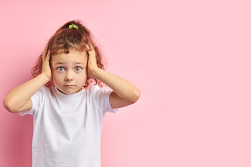 Frightened kid with auburn hair and big eyes, covered ears while standing on pink background