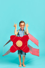 Caucasian boy wearing casual clothes imagine flight in sky on cardboard airplane, in blue sky. Portrait isolated blue background