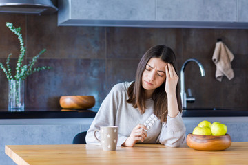 Brunette girl is sitting at table in kitchen and holding hand on head temple. Young woman is feeling bad and going to take painkiller pills. Sudden attack of migraine and headache. Effects of stress.