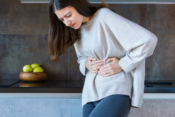 Young woman in grey clothes is holding hands on belly. Brunette girl is feeling bad and sick....