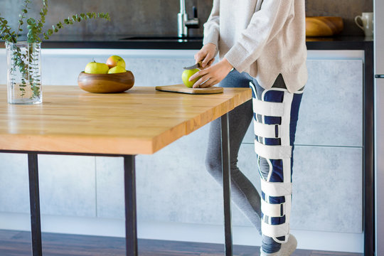 Girl with broken leg is standing in kitchen and cooking. Injured woman wearing supporting compression bandage for trauma to help promote recovery of bones, muscles, ligaments. Orthopedic diseases.