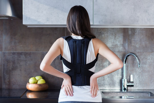 Brunette girl is standing in kitchen and holding hand on back. Young woman dressed orthopedic bandage for relieve spine stress, correct posture, restore after injury. Treatment of spinal diseases