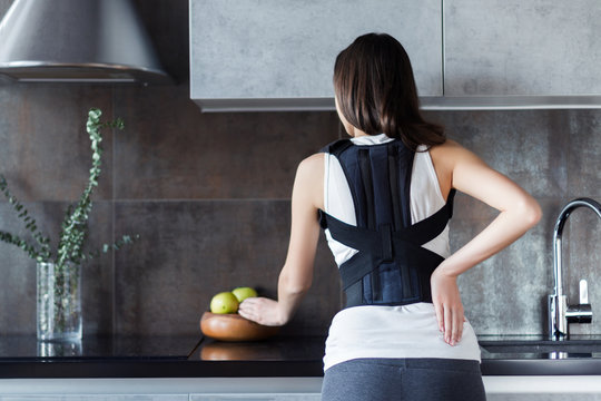 Brunette girl is standing in kitchen and holding hand on back. Young woman dressed orthopedic bandage for relieve spine stress, correct posture, restore after injury. Treatment of spinal diseases
