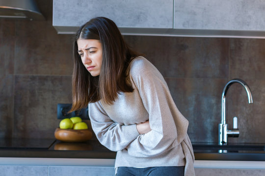 Young woman in grey clothes is holding hands on belly. Brunette girl is feeling bad and sick. Sudden onset of diarrhea, stomach ache, pancreatitis, appendicitis attack. Bad junk food concept.