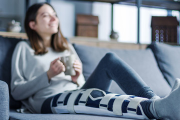 Smiling brunette girl with broken leg is sitting on couch sofa, resting and drinking tea at home....
