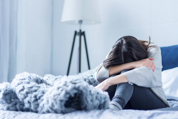 Young woman in gray clothes is sitting curled up on bed at home. Upset brunette girl is suffering, crying. Depression because of loneliness, stress, problems. Psychological disorders concept. - obrazy, fototapety, plakaty