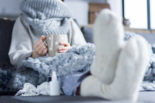 Sick girl in scarf and knitted socks is sitting in bed sofa wrapped in grey blanket. Ill woman is drinking tea and taking pills medications. Home treatment. Winter cold and flu concept.