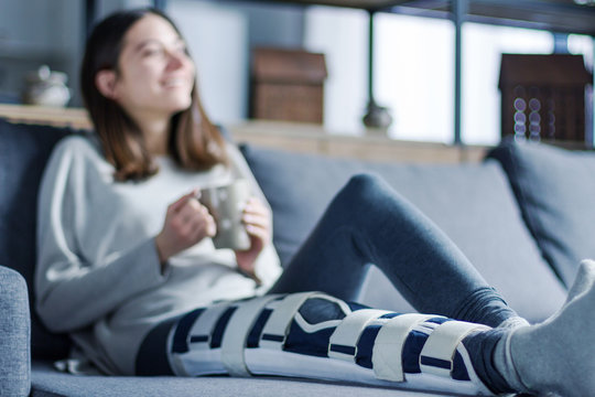 Smiling brunette girl with broken leg is sitting on couch sofa, resting and drinking tea at home. Injured young woman wearing supporting compression bandage for trauma. Sick leave concept.