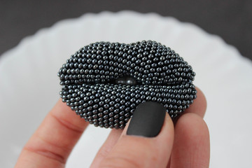 Lips-shaped brooch made of beads in hands with black manicure. Black background with white plate.