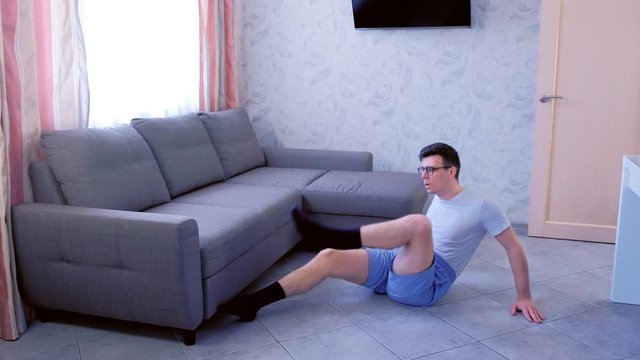 Funny nerd man in glasses and shorts is doing abs exercise lifting his legs to chest at home. Sport humor concept.