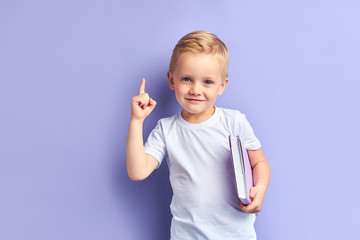 Caucasian kid show bright emotions standing over purple background, holding purple notebook.