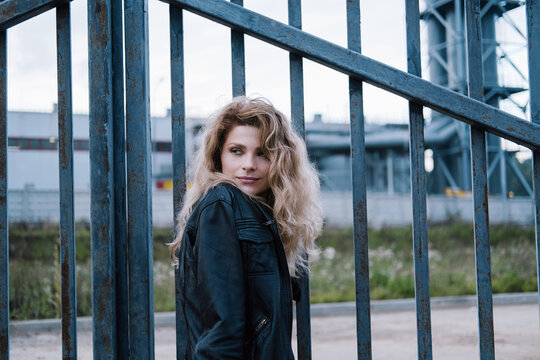 beautiful female wearing leather jacket standing outdoor. iron fence and Industrial landscape on background
