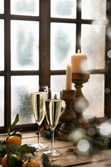 Christmas table at the winter window. Two glasses of champagne, candles in wooden candlesticks and tangerines surrounded by fresh spruce branches