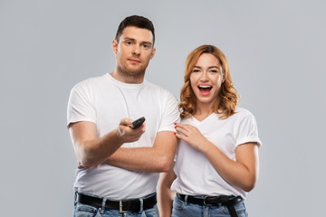 Fototapeta premium entertainment, television and people concept - couple in white t-shirts with tv remote control over grey background