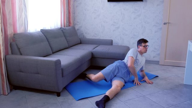 Weak nerd man in glasses is doing funny exercises for legs laying on the floor at home. Sport humor concept.