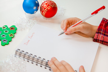 Dear Santa letter, Christmas card. A child holding a pen writes on a white sheet on a wooden background with New Year's decor.