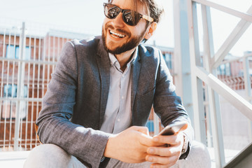 Portrait of happy manager with stubble sitting and using mobile phone outdoors