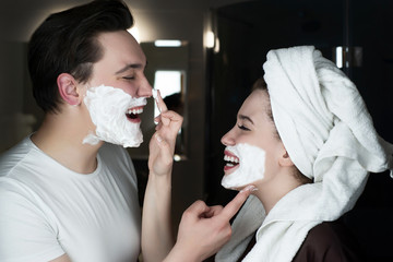 funny couple beautiful woman applying jokingly foam on handsome man's nose having fun both with shaving foam on their faces in bathroom laughing playful mood
