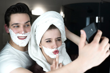funny couple handsome man and beautiful woman striking a pose for selfie looking funny both with shaving foam on their faces in bathroom playful mood