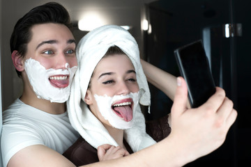 funny couple handsome man and beautiful woman striking a pose for selfie having fun both with shaving foam on their faces in bathroom playful mood