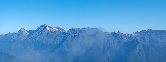 Fototapeta premium Panorama of mountain ranges in sunny early morning; clouds floating on peaks, first sunrise sunbeams; Chugush mount is highest point in area; spruce-fir forests on slopes; higher subalpine meadows