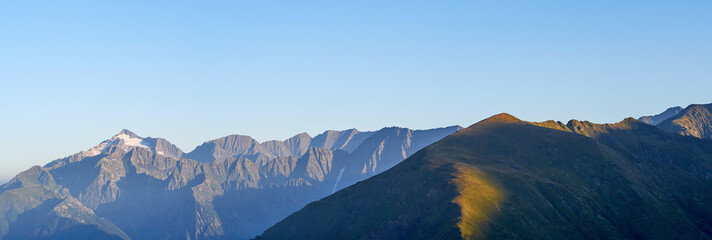 Panorama of mountain ranges in sunny early morning; clouds floating on peaks, first sunrise sunbeams; Chugush mount is highest point in area; spruce-fir forests on slopes; higher subalpine meadows