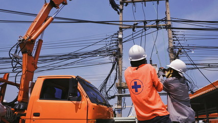 Rear view and selective focus at 2 electrician workers with crane truck on the road are working to installation cable lines on electric power poles with blue sky background