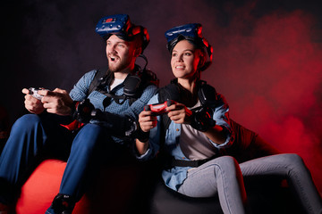 Cheerful couple playing videogame sitting on sofa isolated over black background, neon backlit. Using joysticks, wearing headsets, goggles