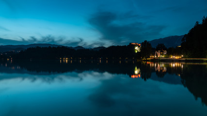 Fototapeta premium Bled lake landscape at dusk, in the blue hour. You can see the hills, Bled Castle, and some clouds; as well as its reflection in the water.