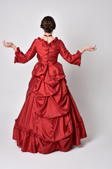 full length portrait of a brunette girl wearing a red silk victorian gown. Standing pose, with back...