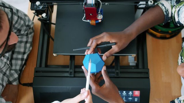 Male designers African American and Caucasian are holding 3d printed shape discussing design while machine is printing plastic element. People and innovation concept.