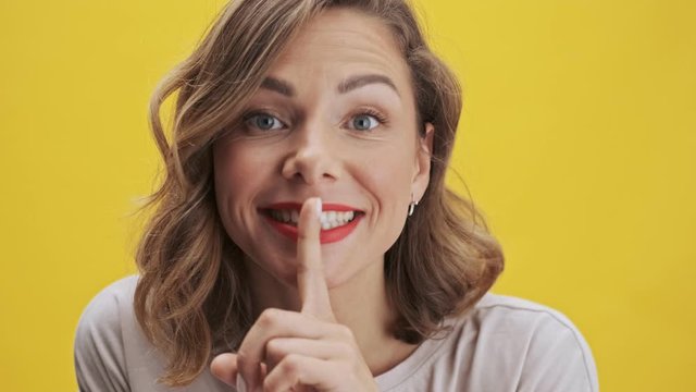 Attactive young woman with red lips making a silence gesture and smiling while looking at the camera over yellow background isolated