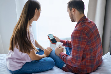 Young pleasant couple sitting on white sheet with backs towards camera, staring at cell phone screen, watching video, trying to call parents, posing against background of light window, indoor shot