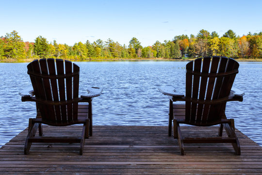 Two Muskoka chairs sitting on a wood dock facing a lake in a calm autumn season sunny day