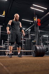 Fototapeta na wymiar Strong brutal bodybuilder dressed in black sports singlet and shorts, wearing sneakers, standing on brown floor of sports studio, getting ready for weight lifting training