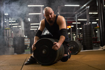 Obraz na płótnie Canvas Young man with bald head, dressed in black stylish sportswear looks aside with interest, sitting on barbell, changing metal weight plate, gym atmosphere, indoor shot