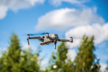 A gray drone flies against the sky and trees.