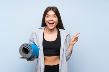 Young sport girl with mat over isolated blue background surprised and pointing finger to the side