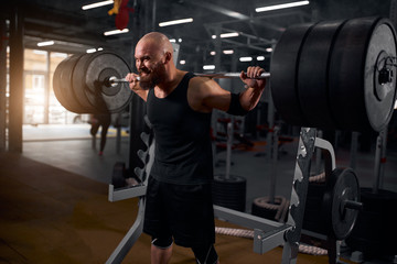 Fototapeta na wymiar Hairless bald powerlifter with thick beard dressed in black sports clothes, attempting to rise heavy barbell, making effort, training hard, preparing for workout, side shot, professional sport concept