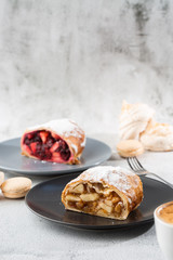 Homemade Traditional Austrian apple strudel with fresh apples, nuts and powdered sugar. Menu for cafe. Piece of cake on black plate, white cup on white marble background. Vertical photo.