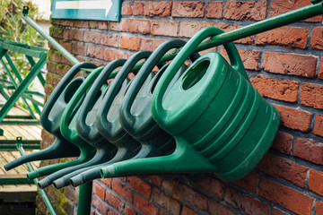 Several watering cans hanging upside down without a spout on a pole on a brick wall with a Wegweiserweg above. In the background a blurred frame